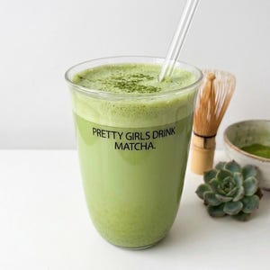Può includere: Un bicchiere di bevanda al matcha verde con una cannuccia di vetro trasparente. Il bicchiere ha il testo "PRETTY GIRLS DRINK MATCHA". Un frullatore per matcha, una piccola ciotola di polvere di matcha e una pianta succulenta sono sullo sfondo.