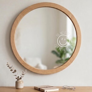 May include: A round wooden framed mirror with a light brown finish. The mirror reflects a room with a plant and the text "DIE WELT IST SCHÖNER, WEIL DU DA BIST" with a smiley face. A small vase with plants, a notebook, and glasses are on a wooden table.
