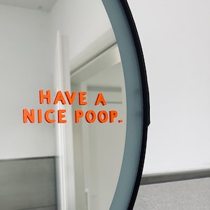 Puede incluir: Espejo redondo con marco negro y texto naranja "HAVE A NICE POOP.". El espejo refleja una pared blanca y una puerta.