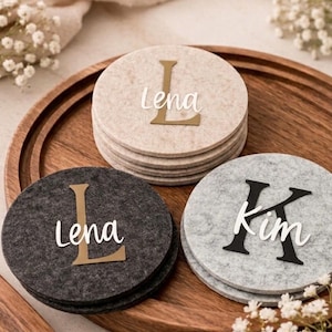 Può includere: Un set di sottobicchieri rotondi in feltro beige, grigio e grigio scuro. Ogni sottobicchiere presenta un nome, come "Lena" o "Kim", con una lettera decorativa. I sottobicchieri sono disposti su un vassoio di legno, con accenti floreali.