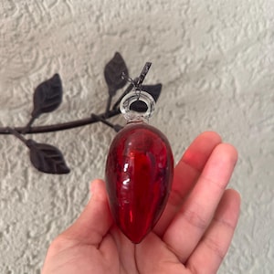 Könnte beinhalten: Ein tropfenförmiges rotes Glasornament mit einer klaren Glasöse an der Oberseite. Das Ornament hat eine glatte, reflektierende Oberfläche und ist an einer dünnen schwarzen Schnur befestigt. Die tiefrote Farbe ist im gesamten Glaskörper einheitlich.