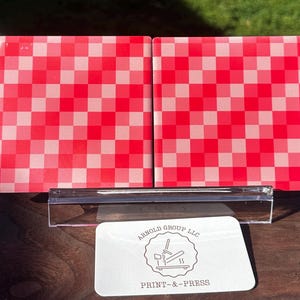 May include: Two square coasters with a red and white checkered pattern. The coasters are displayed on a clear acrylic stand. A white card with the text "ARNOLD GROUP LLC PRINT-&-PRESS" is in front.