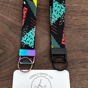 May include: Two wrist keychains with a black background and a colorful geometric pattern. One keychain has a rainbow-colored metal ring and the other has a black metal ring. A white card with the text "ARNOLD GROUP LLC" and "PRINT-&-PRESS" is at the bottom.