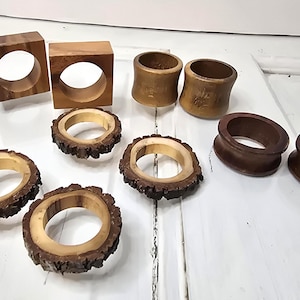 Op de afbeelding: Een verzameling houten servetringen in verschillende stijlen. Inclusief vierkante, cilindrische en schorsrand ontwerpen. De kleuren variëren van licht- tot donkerbruin, wat een rustieke en natuurlijke uitstraling geeft. Perfect voor tafeldecoratie.