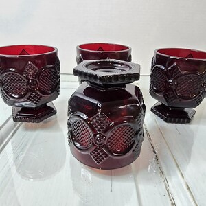 Puede incluir: Un conjunto de cuatro copas de color rojo rubí y un tapón a juego. Las copas tienen un patrón texturizado y se asientan sobre una base cuadrada. El tapón tiene un diseño geométrico. La cristalería es probablemente vintage.