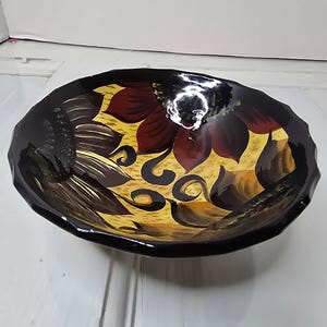 Könnte beinhalten: Eine dekorative, handbemalte Schale mit schwarzem Rand und floralem Design. Der Innenraum hat einen gelben Hintergrund mit braunen und burgunderfarbenen Blumen. Die Schale ist wahrscheinlich aus Keramik oder einem ähnlichen Material.