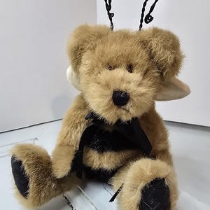 May include: A plush teddy bear with a bee-themed design. The bear has tan fur, a black and yellow striped body, and black paws. It features black wire antennae and a black bow. The bear is sitting down.