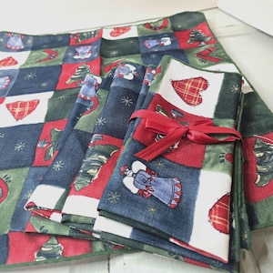 Pode incluir: Um conjunto de guardanapos de tecido com tema natalino e um jogo americano a condizer. Os guardanapos são amarrados com uma fita vermelha. O tecido apresenta um design de patchwork com quadrados vermelhos, verdes, brancos e azuis, decorados com anjos, corações e árvores de Natal.