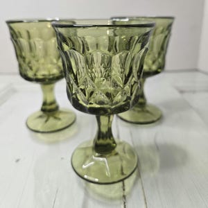May include: Four olive green vintage goblets with diamond-cut patterned bowls and stemmed bases. The glasses are arranged on a white surface, showcasing their intricate design and color. The goblets are ideal for serving beverages.
