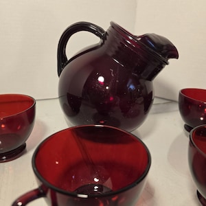 Puede incluir: Una jarra de vidrio rojo rubí vintage con asa y boquilla, acompañada de varias tazas a juego. El juego de cristalería es probablemente para servir bebidas y tiene un diseño clásico y elegante. Las tazas tienen asas pequeñas.