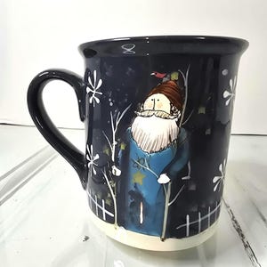 May include: A dark blue ceramic mug with a whimsical winter gnome design. The mug features a white beard, a blue coat, and a brown hat. White snowflakes and a white picket fence are also painted on the mug. The mug has a black handle and a white base.