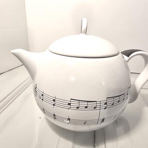 以下が含まれることがあります： 蓋と取っ手が付いた白いセラミックティーポット。ティーポットは、ボディの周りに黒い音符と五線譜で装飾されています。ハンドルには黒いアクセントがあります。ティーポットは白い表面にあります。