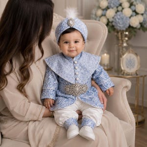 May include: A baby wearing a light blue and white patterned outfit, including a jacket, pants, and a turban-style hat with a feather. The outfit features a decorative silver belt. The baby is sitting on a person's lap.