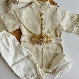 Könnte beinhalten: Elfenbeinfarbenes formelles Outfit mit goldenen Akzenten. Das Set beinhaltet eine Jacke mit goldenen Knöpfen und einem dekorativen Gürtel, eine passende Hose, einen Hut und zwei kleine Schuhüberzieher. Der Stoff hat ein subtiles, verziertes Muster.