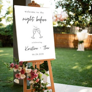 May include: A white welcome sign on a wooden easel with the text "welcome to the night before" and the names "Kristine + Theo" with the date 06.28.2028. The sign is decorated with a floral arrangement of pink and white flowers.
