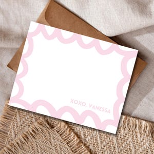 Puede incluir: Una tarjeta de papelería blanca con un borde festoneado rosa claro y el texto "XOXO, Vanessa". La tarjeta está colocada sobre un sobre marrón y una superficie beige texturizada.