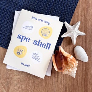 Op de afbeelding: Een wenskaart met de tekst "you are very spe-shell to me!" en illustraties van schelpen. De kaart wordt vergezeld door een witte zeester, een grote bruine schelp en twee kleinere witte schelpen. De items zijn gerangschikt op een houten oppervlak.