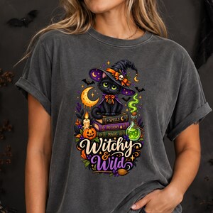 May include: A charcoal gray short-sleeve t-shirt featuring a graphic of a black cat wearing a witch hat, sitting on a stack of books labeled Spells, Potions, and Magic. The design includes a crescent moon, a candle, a pumpkin, and the text Witchy & Wild.