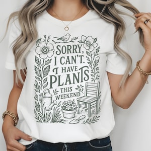 May include: White t-shirt with a green and white graphic that reads "Sorry, I can't. I have plants this weekend." The graphic includes illustrations of flowers, a watering can, a chair, and a trowel. The shirt is a crew neck.