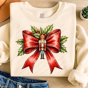 May include: Cream-colored sweatshirt featuring a festive design. A large red bow with white stitching is the focal point, adorned with a detailed nutcracker figure and holly leaves. The design evokes a holiday or Christmas theme.