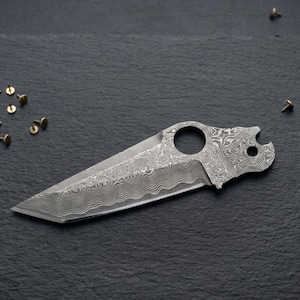 May include: A detailed, patterned metal knife with a sharp blade, a circular hole, and a unique handle design. Small, brass-colored tacks are scattered around the knife on a dark surface. The knife's blade has a wavy edge.