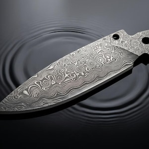 Könnte beinhalten: Eine Messerklinge aus Damaszenerstahl mit einem ausgeprägten, wirbelnden Schichtenmuster in Grau- und Silbertönen. Die Klinge hat eine spitze Form und einen Erl mit zwei kreisförmigen Löchern zur Befestigung eines Griffs.