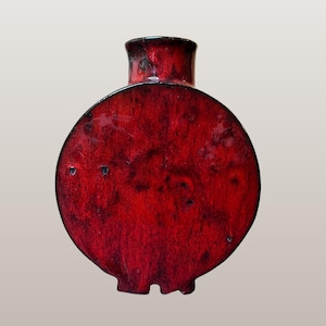 May include: A red, circular vase with a short neck and a flat base. The vase has a textured surface and a dark rim. The text "WGP207 Studio Pottery | Red Circle Vase" is at the bottom of the image.