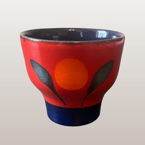 Può includere: Una tazza da punch o fioriera in ceramica rossa con base e bordo blu. La tazza presenta un cerchio arancione e disegni neri a forma di foglia. Il testo "WGP339" e "9cm Tall" sono visibili.