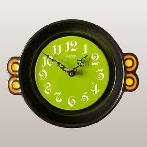Peut inclure: Une horloge murale ronde avec un cadran vert et des chiffres blancs. L'horloge a un cadre noir avec des accents orange et jaune sur les côtés. Le texte "Pallas Heges Wall Clock | 3122 | Green" est visible.
