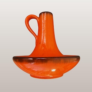 Puede incluir: Candelabro de cerámica naranja con asa y base ancha y poco profunda. El candelabro tiene un acabado brillante y un borde marrón oscuro. El texto "WGP259", "Dümler & Breiden 517/14 | Candleholder" está en la parte inferior.