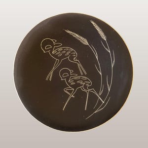 Può includere: Un piatto da parete rotondo, marrone scuro, con un disegno di cervo. Il piatto presenta una tecnica a sgraffito con un cervo e spighe di grano incise in un colore più chiaro. Il piatto ha un diametro di circa 20 cm.