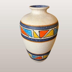 Puede incluir: Un jarrón de cerámica color crema con superficie texturizada. Presenta un borde azul y una banda de formas geométricas azules, naranjas y amarillas. El texto "WGP196 Ü-Keramik "Memphis" 1001/31" es visible.