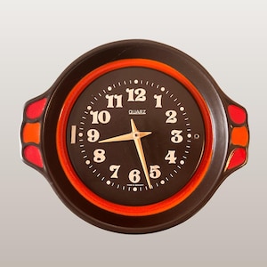 Op de afbeelding: Een ronde, donkerbruine quartz wandklok met een zwarte wijzerplaat en witte cijfers. De klok heeft een rode binnenring en decoratieve rode en oranje accenten aan de zijkanten. De tekst "QUARZ" is zichtbaar. De tekst "WGP355 Pottery Wall Clock | H 3330" staat onderaan.