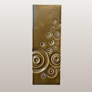 Può includere: Una piastrella rettangolare in ceramica color bronzo con un motivo di cerchi concentrici. I cerchi variano di dimensioni e sono disposti a cascata. La piastrella è di Celtic Ceramics, Irlanda.
