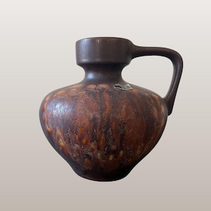 Może przedstawiać: Brązowy ceramiczny dzban z uchwytem, ​​o teksturowanej powierzchni z odcieniami brązu i pomarańczy. Dzban ma szeroką podstawę i wąską szyjkę. Widoczny jest tekst "WGP142 Silberdistel 306-16".