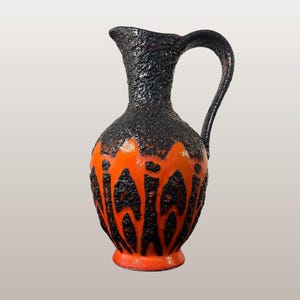 Può includere: Brocca in ceramica con una parte superiore nera strutturata e una parte inferiore arancione vibrante. La sezione arancione presenta un motivo nero audace e astratto. La brocca ha un manico e un beccuccio. Il testo "WGP140 ES Keramik "Jaguar" 880/30" è visibile.