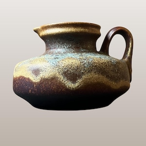 Może przedstawiać: Ceramiczny dzban z uchwytem i wylewką. Dzban ma ciemnobrązową podstawę, która przechodzi w jaśniejszą brązową i złotą krawędź. Korpus ma niebiesko-złoty pas. Tekst "WGP183 Silberdistel | 347/12" znajduje się na dole.