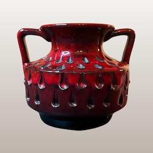 Op de afbeelding: Een rode keramische vaas met twee handvatten en een getextureerd oppervlak. De vaas heeft druppelvormige inkepingen en zilveren accenten. De basis is zwart. De vaas is een decoratief item.