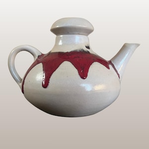 Può includere: Una teiera in ceramica bianca con corpo arrotondato, manico curvo e beccuccio. La teiera è decorata con un audace motivo a smalto rosso gocciolante. Il coperchio è bianco e arrotondato. Testo sull'immagine: WGP309, Elke & Elmar KUBICEK | 144/1 | Teapot.