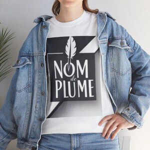 May include: White t-shirt with a black and white graphic design featuring a feather and the words "NOM de PLUME". The shirt is worn with a denim jacket and blue jeans. The design is a modern, minimalist style.