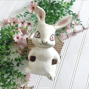 May include: A white ceramic bunny figurine with pink accents on the ears and eyes, holding a small wooden cart. The bunny has a smiling expression and is surrounded by pink flowers and green foliage, creating a spring or Easter theme.