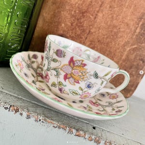 May include: A white teacup and saucer with a floral pattern in shades of pink, yellow, green, and red. The cup and saucer have a green trim.