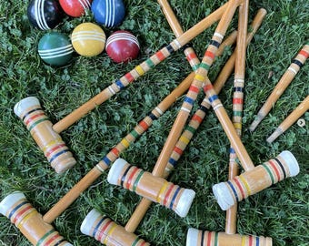 May include: A croquet set with wooden mallets and colorful balls. The mallets have striped handles and white mallet heads. The balls are red, blue, yellow, green, and black with white stripes. The set is arranged on green grass.
