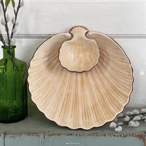 May include: A two-piece ceramic serving dish set in the shape of a seashell. The larger dish is a light beige with brown trim, and the smaller dish sits inside. The dishes have a ribbed texture.