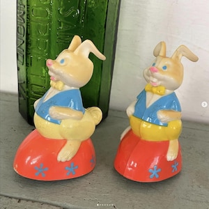 May include: Two vintage Easter bunny figurines. The bunnies have tan fur, blue eyes, and pink noses. They wear blue jackets and yellow bow ties. The bunnies sit on red bases decorated with blue flower shapes. A green bottle is in the background.