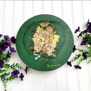 May include: A round, green metal tin with a floral bouquet design. The bouquet features pink and yellow roses tied with a white ribbon. The tin has the text "Drury Lane New York" in a script font.