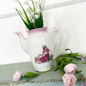 May include: White porcelain pitcher with a pink rim and handle, decorated with a vintage illustration of a girl holding a baby. The pitcher is filled with white flowers and green stems. Pink roses and greenery are placed near the pitcher.