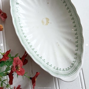 May include: An oval ceramic serving dish with a scalloped edge and a white base. The interior features a green printed border pattern of small clover-like motifs and a single light brown circular stain near the center of the basin.