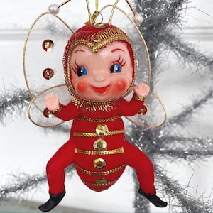 May include: A red and gold Christmas ornament shaped like a bee with a smiling face and wings. The bee is wearing a red and gold costume with gold trim and sequins.