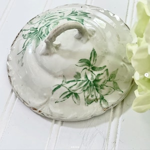 May include: A white ceramic lid with a green floral design and a curved handle. The lid has a scalloped edge with gold accents. The floral pattern features green leaves and flowers. The lid is likely for a decorative container.